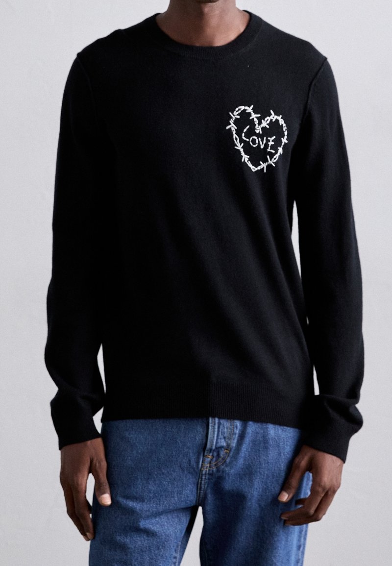 Homme portant un pull noir à manches longues avec un cœur en fil barbelé blanc et le texte « LOVE » sur la poitrine, accompagné d'un jean bleu.