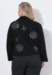 Femme aux cheveux blonds portant une veste noire ornée de motifs de flocons de neige en sequins argentés et un pantalon noir à rayures, faisant face à un mur blanc.