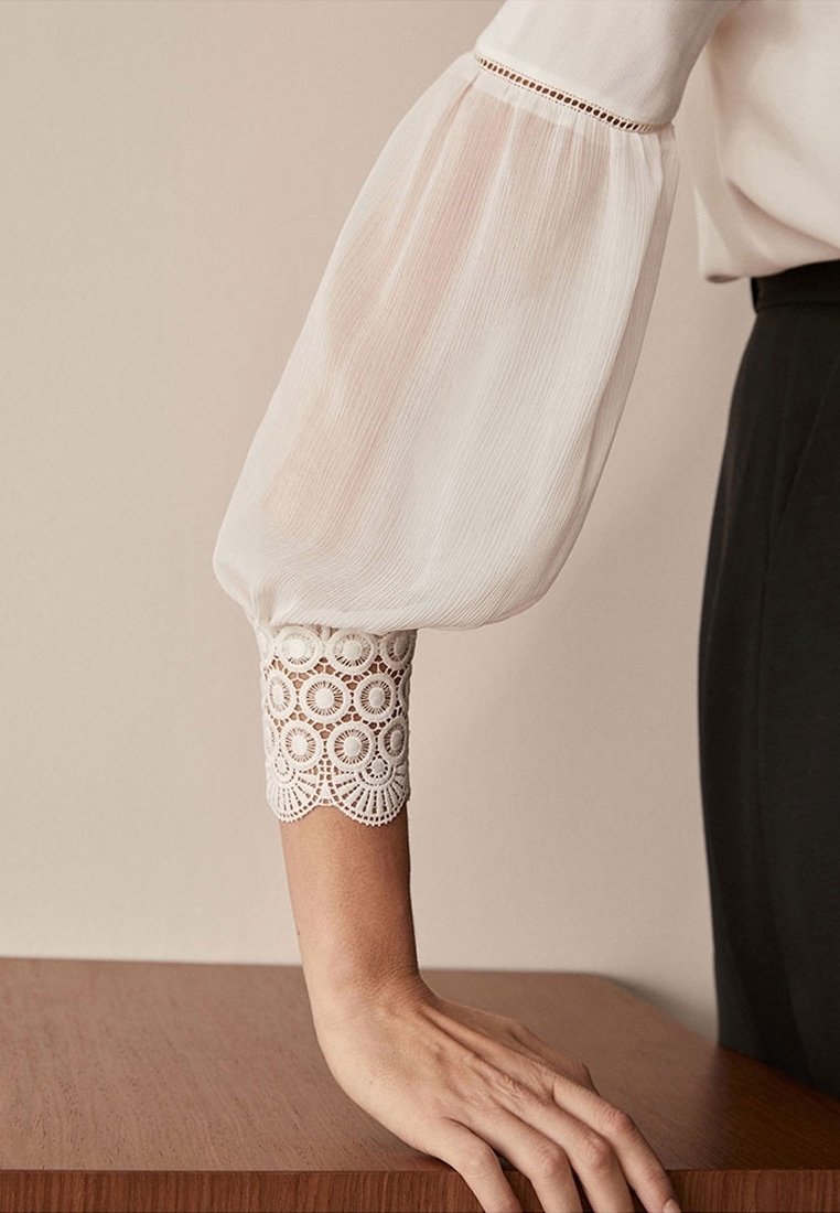 Blouse blanche avec des manches amples et transparentes, et des détails en dentelle aux poignets. Tissu lisse avec une texture légère et aérée. Main reposant sur une surface en bois.