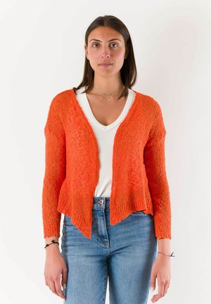 Donna con capelli lisci e scuri indossa un cardigan arancione a maglia aperta sopra una maglietta bianca con scollo a V e jeans blu, in piedi davanti a uno sfondo bianco.