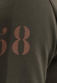 Olívovo zelená tkanina obsahuje veľký hnedý potlač "58". Textúra je ryhovaná, s detailným pohľadom na zaoblené tvary čísla.
