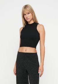 Ung kvinde med glat, blond hår iført en ærmeløs sort crop top og mørke grå høj talje bukser foran en ensfarvet hvid baggrund.