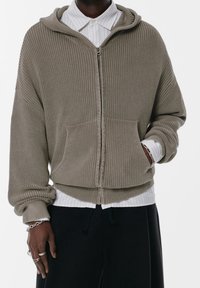 Sweat à capuche zippé kaki en maille côtelée, avec une grande poche avant, une coupe décontractée et des poignets retroussés. Porté sur une chemise blanche à rayures.