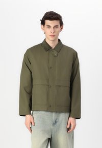 Olive green jacket with a snap closure, corduroy collar, and two large front pockets; made of sturdy fabric with a smooth texture.