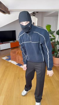 Casaco desportivo preto com fecho de correr, apresentando detalhes em branco. Calças pretas de corte ajustado e ténis prateados. Ambiente interior com plantas.