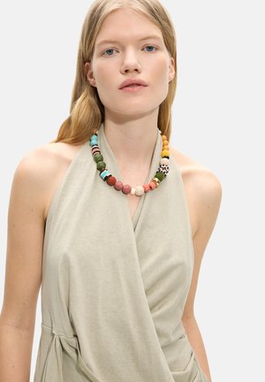 Blondynka w beżowej bezrękawnikowej bluzce halter i kolorowym naszyjniku z koralików, na jednolitym jasnym tle.