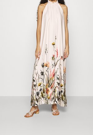 Vrouw in een lichtroze halternek maxi-jurk met een kleurrijke bloemenprint aan de zoom en witte sandalen met straps.