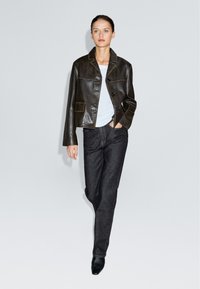 Giacca cropped in pelle marrone con chiusura a bottone e due tasche, abbinata a jeans in denim scuri e stivaletti neri con tacco.