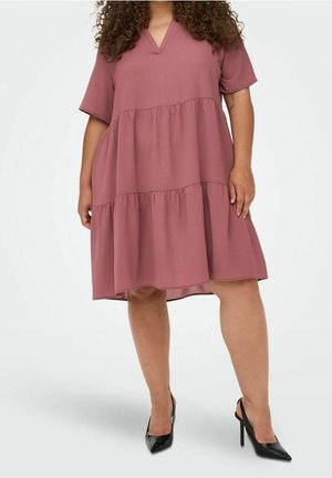 Mujer con pelo rizado, vestida con un vestido suelto de varios volantes en tono rosa empolvado y zapatos negros de tacón alto, de pie frente a un fondo liso.