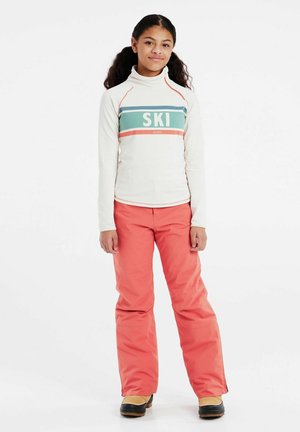 Langärmlige, off-white Skijacke mit türkisfarbenen und korallenfarbenen Streifen, versehen mit dem Text "SKI". Kombiniert mit leuchtend korallenfarbenen Schneehosen und schwarzen Stiefeln.