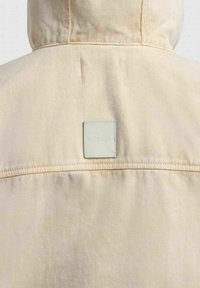 Veste beige à capuche vue de dos avec une couture horizontale cousue et une petite étiquette carrée en tissu centrée sous la capuche.