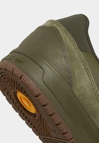 Zapatilla verde con una parte superior texturizada de materiales de ante y sintéticos, suela de goma con un detalle circular naranja y costuras reforzadas.