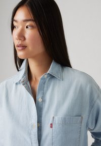 Giovane donna con lunghi capelli scuri che indossa una camicia in denim azzurro chiaro con bottoni, con una tasca sul petto visibile e etichetta rossa Levi's, che guarda verso sinistra.