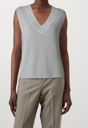 Personne à la peau foncée portant un haut sans manches en maille gris clair à col en V et un pantalon beige sur un fond uni.