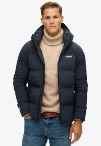 Marineblå puffjakke med hætte, lynlåslukning og logo detalje, båret over en beige strikket turtleneck. Casual denimjeans og et brunt bælte.