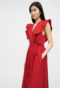 Γυναίκα που φοράει κόκκινο φόρεμα χωρίς μανίκια με βολάν στους ώμους και ζώνη στη μέση, στέκεται μπροστά σε ένα απλό φωτεινό φόντο.