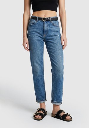 Vrouw draagt opgerolde blauwe spijkerbroek, zwarte riem, zwart cropped top en zwarte gespen sandalen, staand op een effen lichte achtergrond.