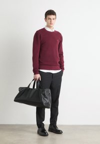 Svart skinn duffelbag med to håndtak, glatt tekstur og lang form. Modellen har på seg en bordeaux genser over en krage skjorte og mørke bukser.