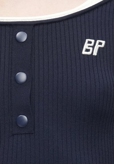 Μπλε ναυτικό ριμπ τοπ με τρία μεγάλα κουμπιά και λευκό κεντητό λογότυπο "BP" στην αριστερή πλευρά κοντά στη γραμμή του λαιμού.