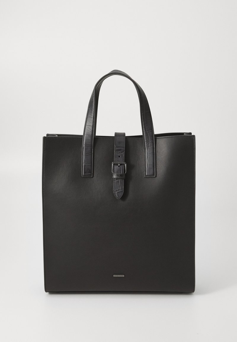 Μαύρη δερμάτινη τσάντα tote με δύο λαβές στην κορυφή και κεντρικό κλείσιμο με λουρί και αγκράφα σε λευκό φόντο.