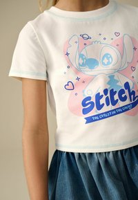 Vit bomullströja med korta ärmar, som har en blå och rosa grafisk bild av en karaktär och texten "stitch" i fet blå skrift.