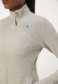 Lys grå zip-jakke med høy krage, med teksturert stoff, kremfargede detaljer, og en liten blå logo på skulderen.