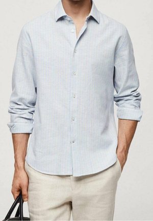 Homme portant une chemise bleu clair à boutons avec les manches retroussées, pantalon beige, tenant un sac noir dans une main et l'autre main dans la poche.