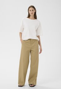 Femme debout, les mains dans les poches, portant un haut blanc ample à manches courtes, un pantalon large beige et des sandales noires sur fond blanc.