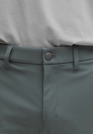Chemise grise portée au-dessus d'un pantalon vert foncé, présentant une texture lisse, une fermeture à boutons et des passants de ceinture, avec une coupe ajustée et des coutures nettes.