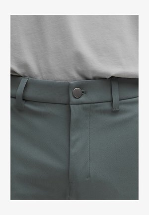 Maglietta grigia sopra pantaloni verde scuro, con una texture liscia, chiusura con bottoni e passanti per la cintura, vestibilità sagomata e cuciture pulite.