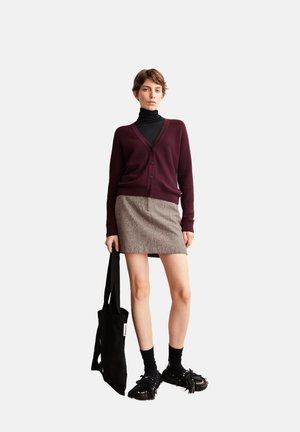 Burgunderfarbener Cardigan über einem schwarzen Rollkragenpullover, kombiniert mit einem strukturierten braunen Rock. Eine schwarze Tote-Tasche und fransige schwarze Schuhe runden den Look ab.