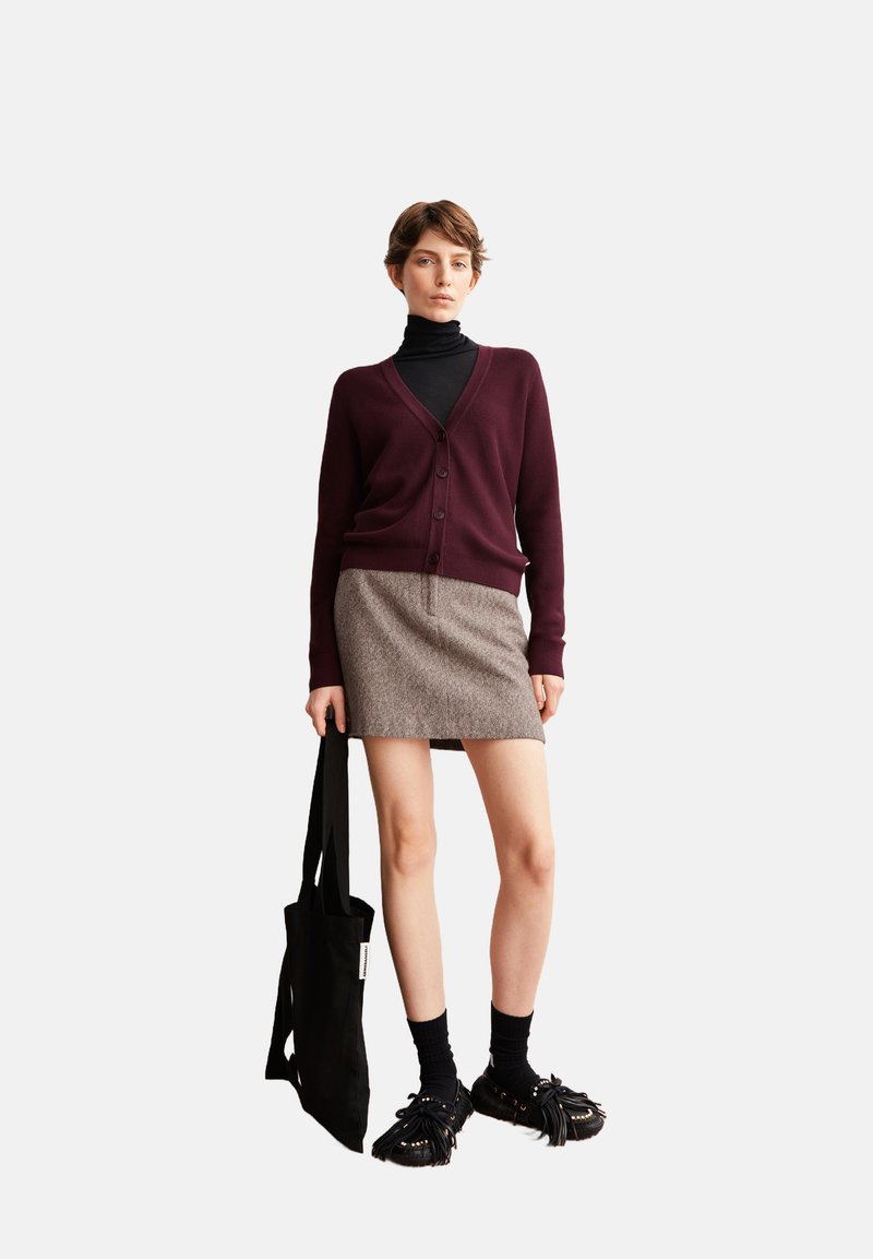Burgunderfarbener Cardigan über einem schwarzen Rollkragenpullover, kombiniert mit einem strukturierten braunen Rock. Eine schwarze Tote-Tasche und fransige schwarze Schuhe runden den Look ab.