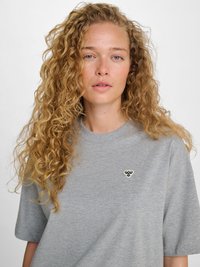 Jeune femme aux longs cheveux blonds bouclés portant un t-shirt gris clair à col rond avec un petit logo d'abeille sur la poitrine, sur un fond uni.