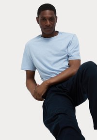 Camiseta de algodón azul claro con cuello redondo, mangas cortas y corte relajado. Combinada con pantalones oscuros de color marino, destacando la posición sentada.