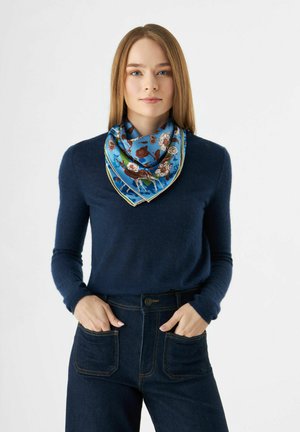 Écharpe en soie à motif floral bleu avec une bordure crème, drapée autour du cou. Portée avec un pull marine et un pantalon en denim foncé.