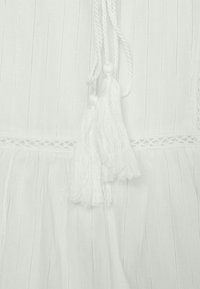 Tissu blanc à rayures verticales avec une surface texturée. Des pompons décoratifs blancs pendent d'un cordon tressé au niveau de l'encolure.