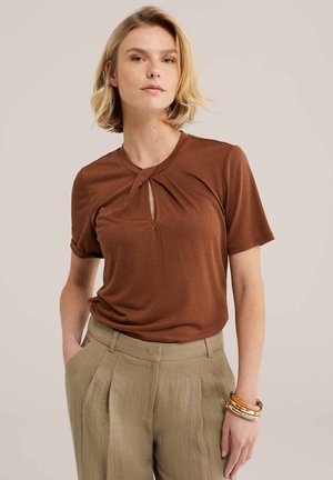 Jeune femme aux cheveux blonds courts portant un haut marron à manches courtes avec un décolleté en trou de serrure et un pantalon beige plissé taille haute.