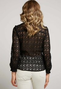 Blouse en dentelle noire avec un motif floral, manches longues et poignets froncés. Texture transparente et taille ajustée, associée à un pantalon de couleur claire.