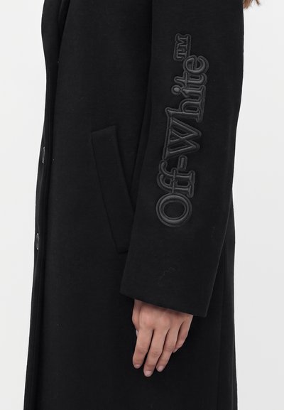 Manteau long noir avec le texte Off-White embossé sur la manche et une main suspendue à côté de la poche latérale.