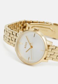 Montre en métal doré avec un cadran rond minimaliste, un compteur blanc et de fins index horaires. Elle dispose d'un bracelet à maillons avec une fermeture à clip.