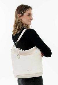 Sac à main en cuir blanc avec une finition douce et texturée. Il dispose d'une grande ouverture, d'une bandoulière ajustable et d'un accent en anneau métallique.
