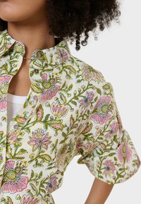 Camicia bianca con bottoni, decorata con un motivo floreale verde e rosa. Presenta un colletto a punta e maniche arrotolate. Tessuto testurizzato con dettagli di cucitura visibili.