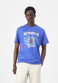 Blaue Baumwoll-T-Shirt mit einem grafischen Design von Wolkenkratzern und einem Superhelden, mit dem Aufdruck "METROPOLIS" und "Superman" in fetten Buchstaben.