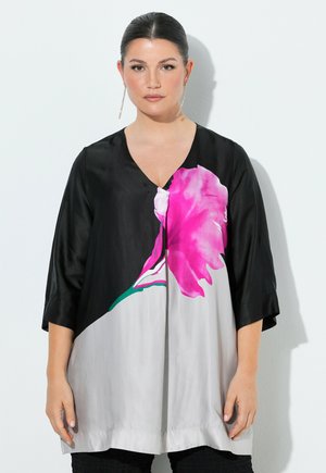 Femme aux cheveux foncés tirés en arrière, portant une tunique noire et grise avec un grand imprimé fleur rose, debout devant un fond blanc.