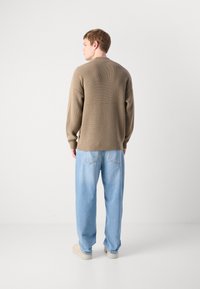 Man facing away wearing a beige knit sweater, light blue jeans, and white sneakers against a plain light background.