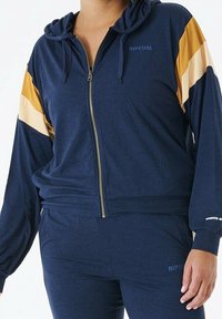 Hoodie zippé marine avec des rayures dorées et crème sur les manches, fabriqué en tissu doux, avec une capuche à cordon et un logo sur la poitrine.