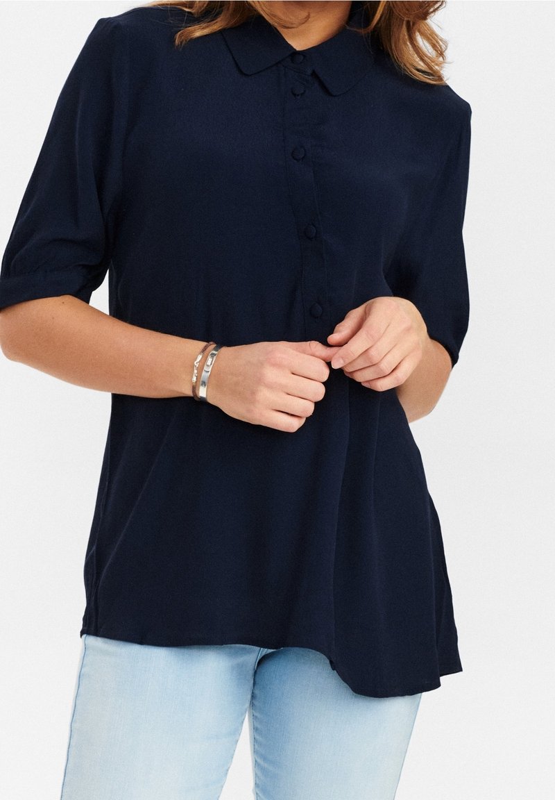 Blusa blu navy con bottoni, colletto, maniche corte e tessuto morbido. Presenta un'orlo curvo e dettagli di bottoni discreti.