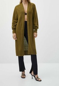 Kvinna klädd i en lång olivgrön öppen cardigan, svarta byxor med framspalter, svarta sandaler med klack och remmar, samt en svart spets-bh.