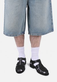 Sandales en cuir noir avec un design tressé, bout ouvert, et sangle de cheville, portées avec des chaussettes blanches de style crew et un short en denim bleu.