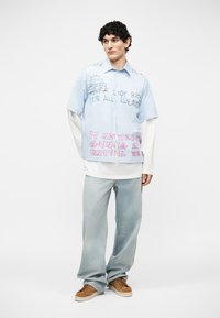 Jeune homme portant une chemise rayée bleue à plusieurs couches avec un texte manuscrit et un jean bleu clair à jambes larges, debout sur un fond blanc.